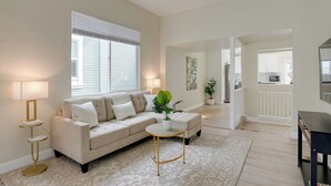 A living room with a beige sofa, a round coffee table, and a lamp. There is a window with blinds, a plant, and a kitchen visible in the background.