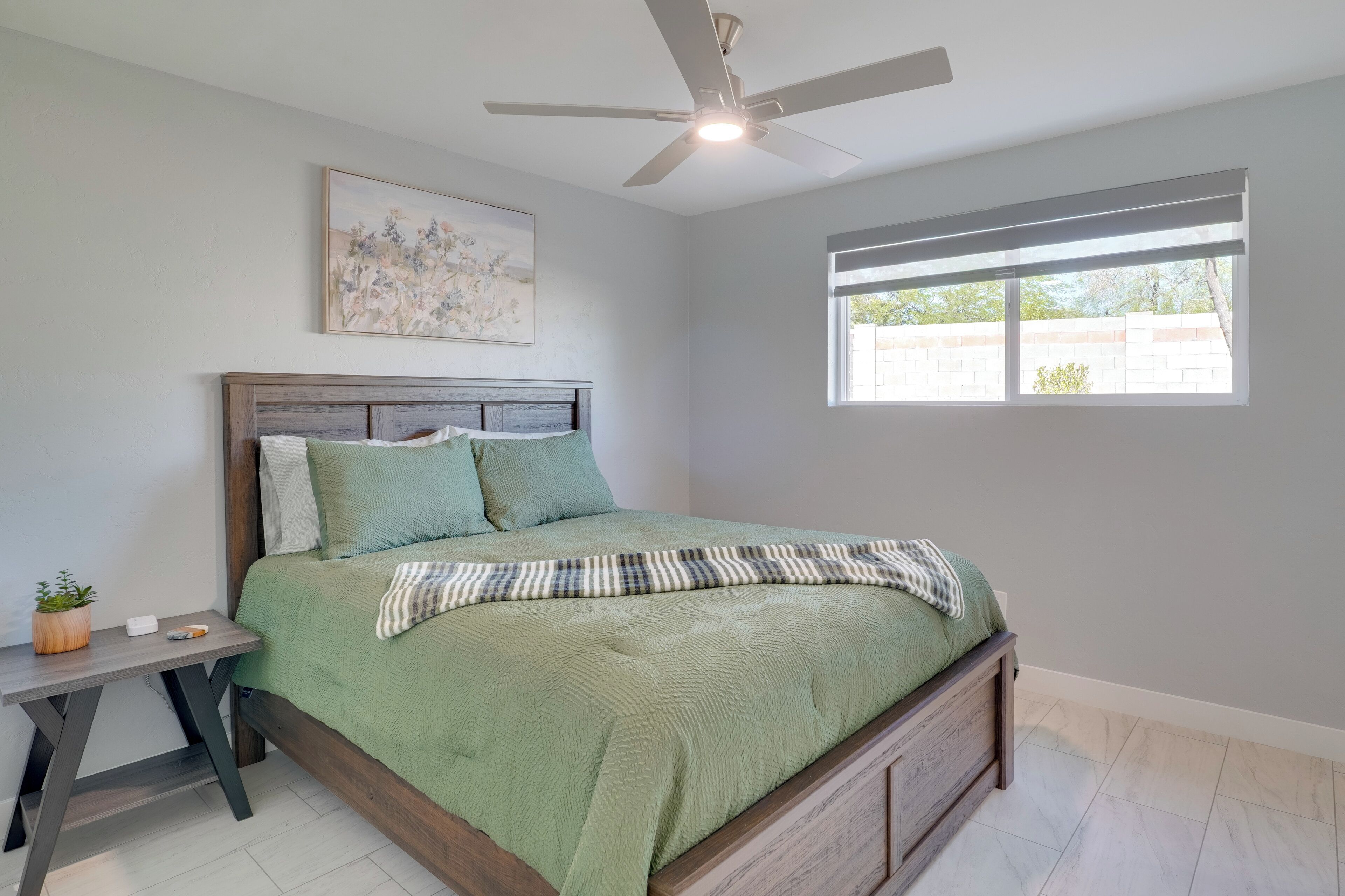 Une chambre à coucher avec un lit, une table de chevet, un ventilateur de plafond, une fenêtre et un tableau au mur.