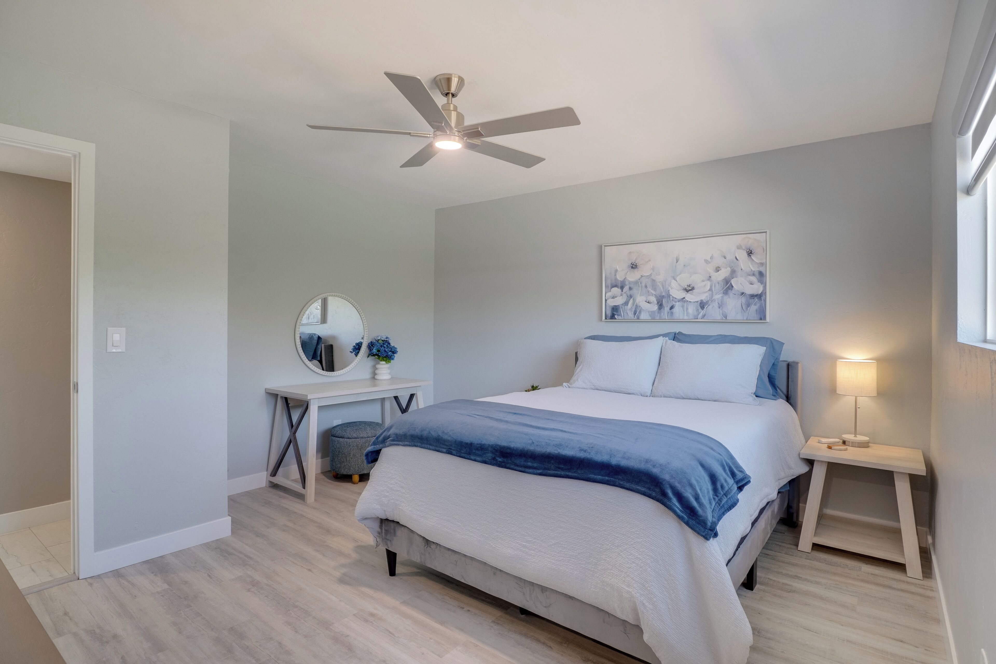 Quarto com cama, escrivaninha, cadeira, ventoinha no teto e mesa de cabeceira com abajur.