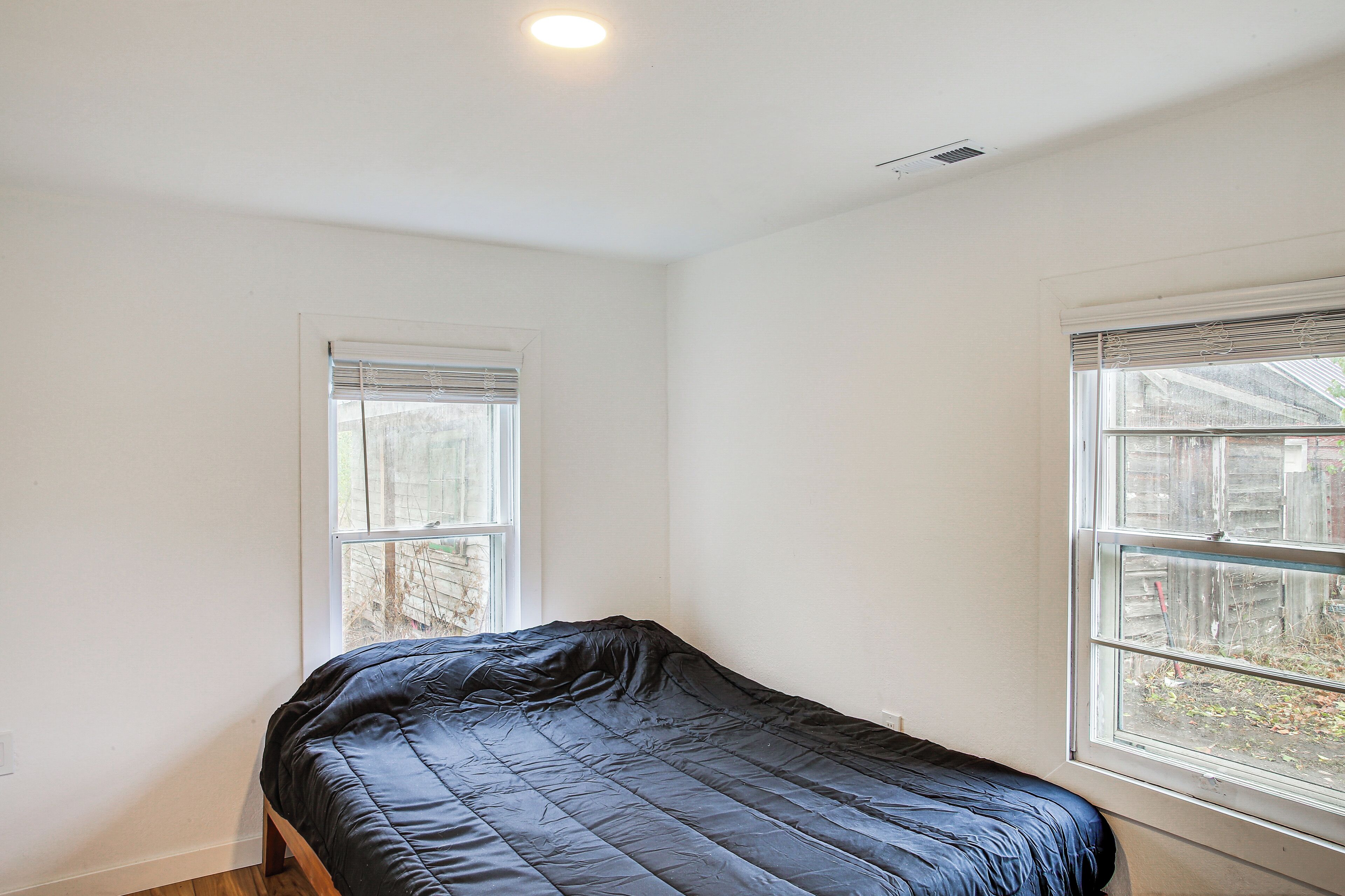 A single bed in a room with two windows, one with blinds and one with a view of the outside.