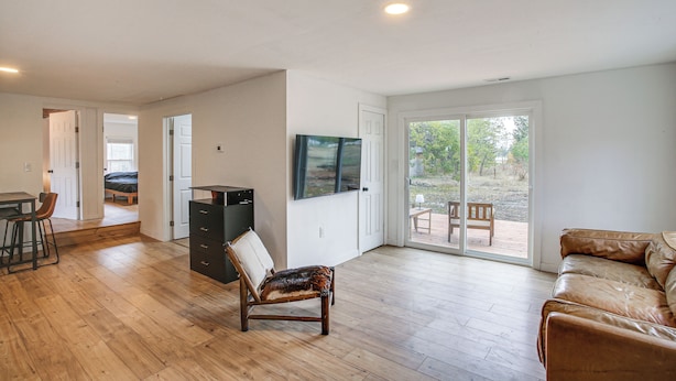 A spacious living room with wooden flooring, a leather sofa, a dining area with a table and chairs, and a sliding glass door leading to an outdoor patio. - Spokane Valley Nature Home! Trails & River Days (Otis Orchards-East Farms)