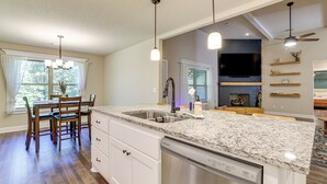 A modern kitchen with granite countertops, a dishwasher, a dining area with wooden chairs, and a built-in TV above the fireplace. - Riders Retreat! Modern Home By Bella Vista Trails (Bella Vista)