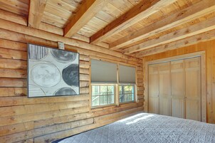Un dormitorio de cabaña de madera con cama, ventana con persianas, puerta y una obra de arte colgada en la pared.