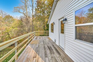 Um deck de madeira com guarda-corpo, um edifício branco com janelas amplas e um fundo arborizado.