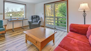 A room with a wooden coffee table, a red sofa, a grey armchair, a small table with two lamps, and a large window with a view of trees. - Dirt Road Digs - Quiet Home in Lakeville! (Lakeville)