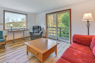 Quarto com mesa de centro de madeira, sofá vermelho, poltrona cinza, mesa pequena com duas luminárias e uma grande janela com vista para árvores.