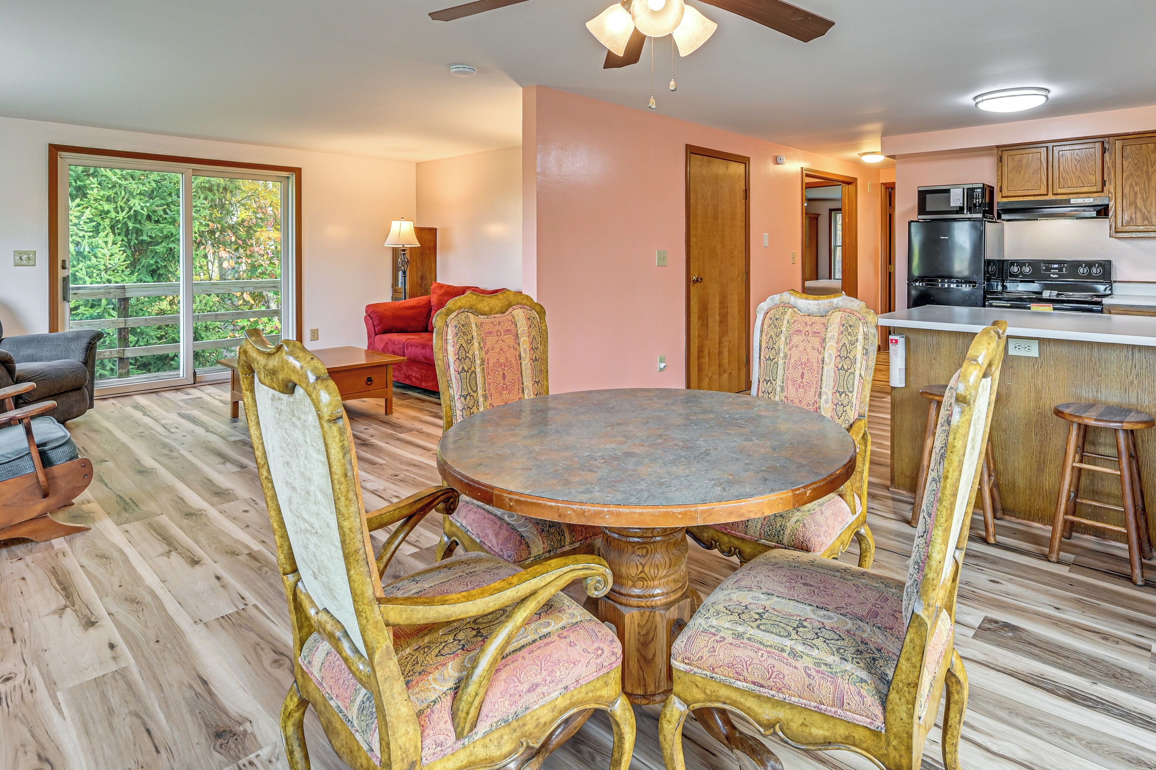 A dining area with a round table and ornate chairs, a kitchen with modern appliances, and a large window with a view of trees.