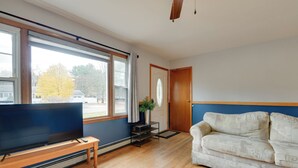 A living room with a TV on a wooden stand, a white sofa, a wooden coffee table, and a large window with a view of a tree and a car outside. - 15 Mi to Cochran's Ski Area: Cozy Home w/ Yard! (Essex Junction)