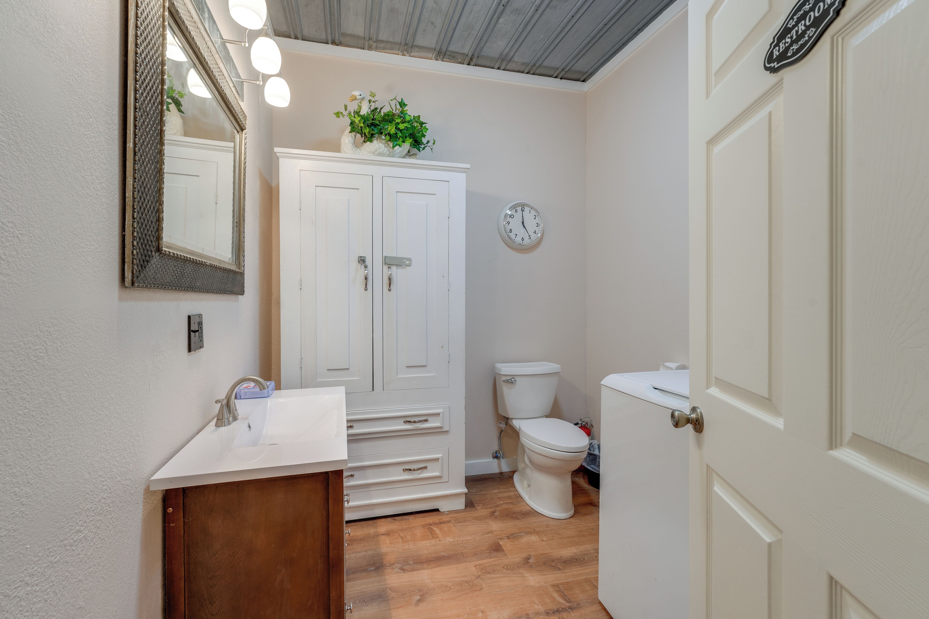Ein Badezimmer mit einem weißen Waschbecken, einem Spiegel, einem Schrank, einer Toilette und einer Uhr an der Wand.