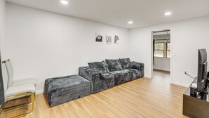 A modern living room with a grey sofa, a white armchair, and a matching ottoman. There is a flat-screen TV on a stand, and the room has white walls and wooden flooring. - Updated Getaway w/ Sauna & Yard in Bloomsbury! (Bloomsbury)