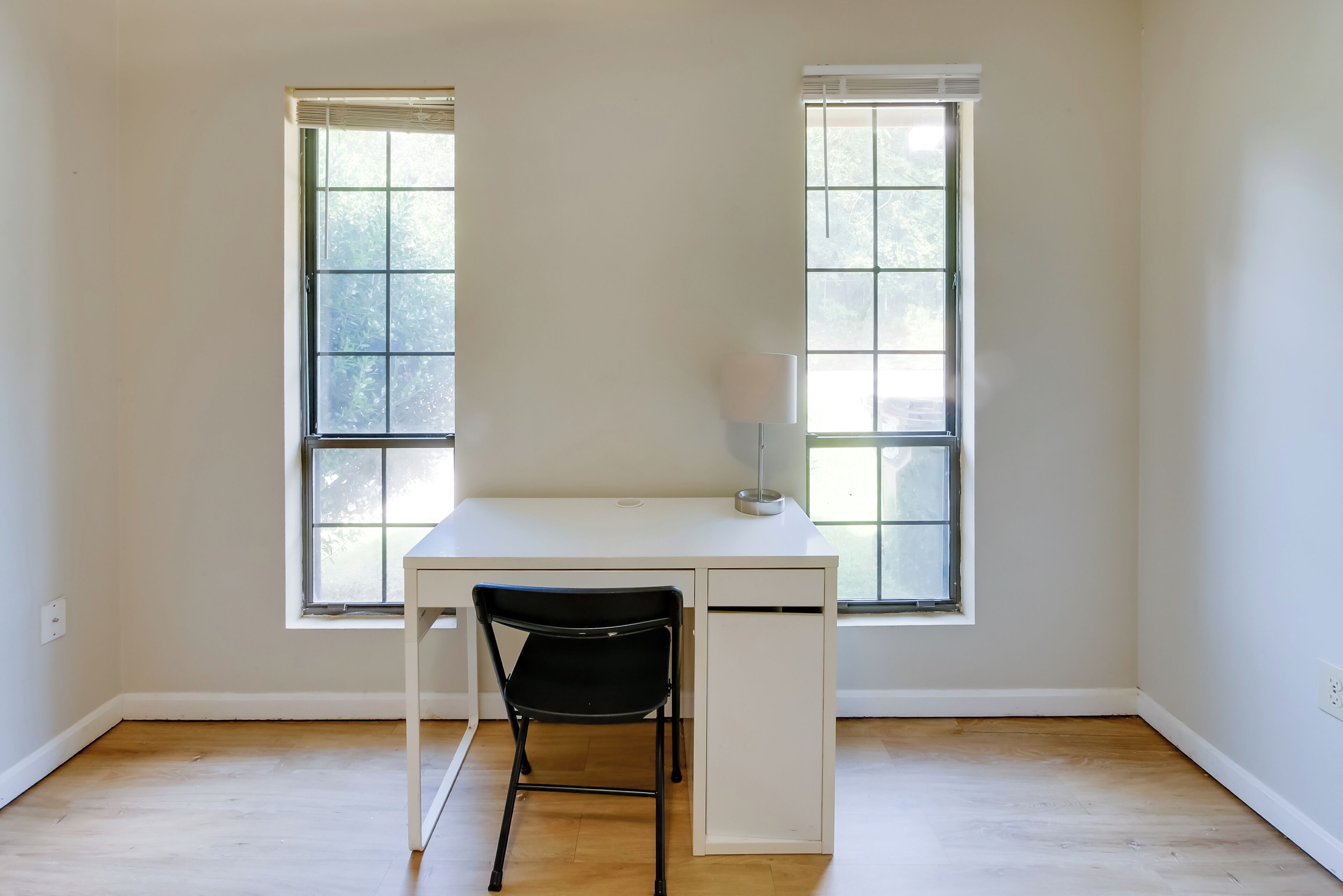 A room with a white desk, two windows, a black chair, and a floor lamp.