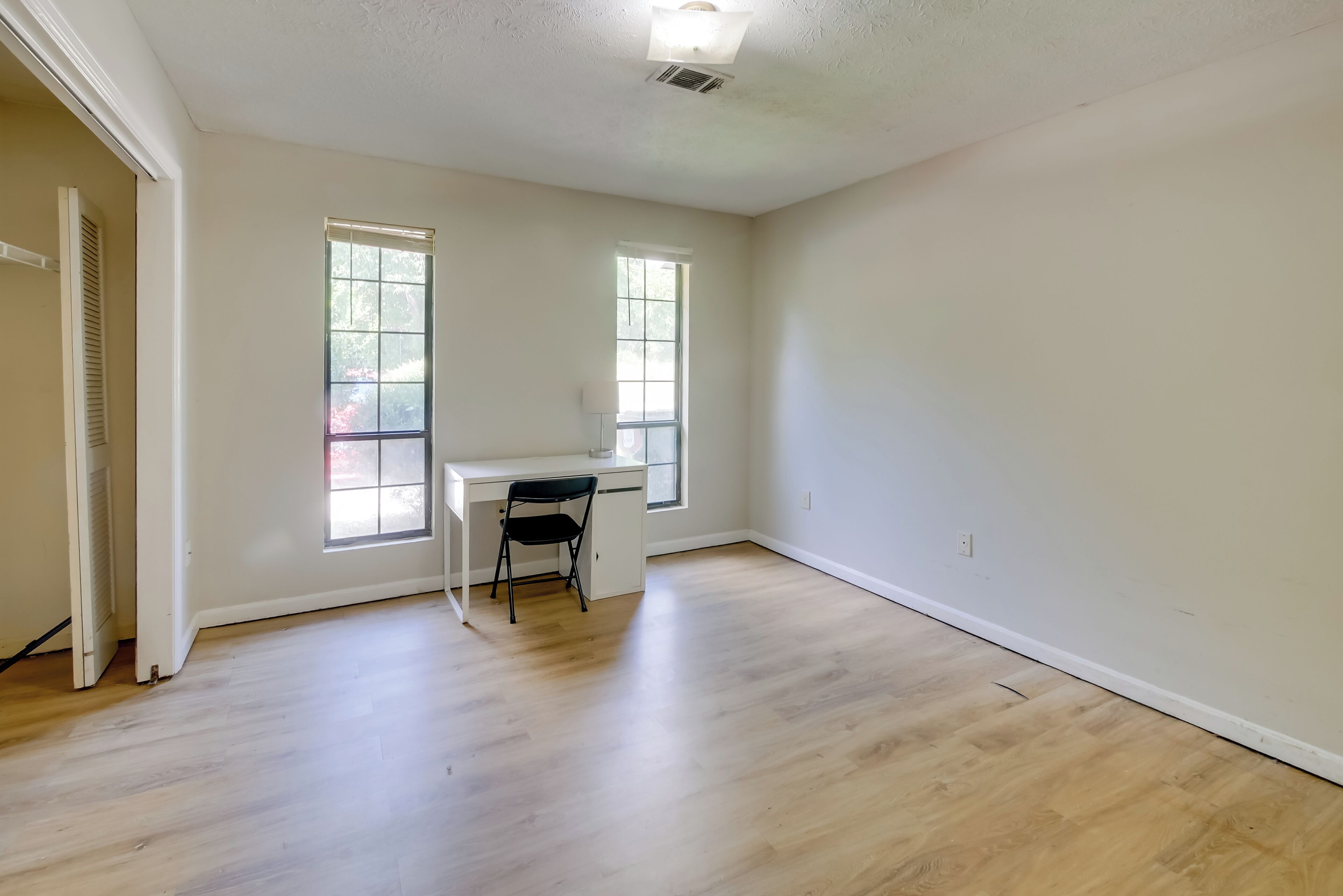 A spacious room with hardwood flooring, a desk with a chair, and two windows providing natural light.