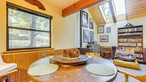 A cozy room with a wooden dining table, a bookshelf, and a large window overlooking a forest. - 1 Mi to Skiing! Cabin Hideaway on June Lake Loop (June Lake)