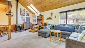 A cozy living room with a wooden ceiling, a fireplace, a bookshelf, and a large window offering a view of the outside. - 1 Mi to Skiing! Cabin Hideaway on June Lake Loop (June Lake)