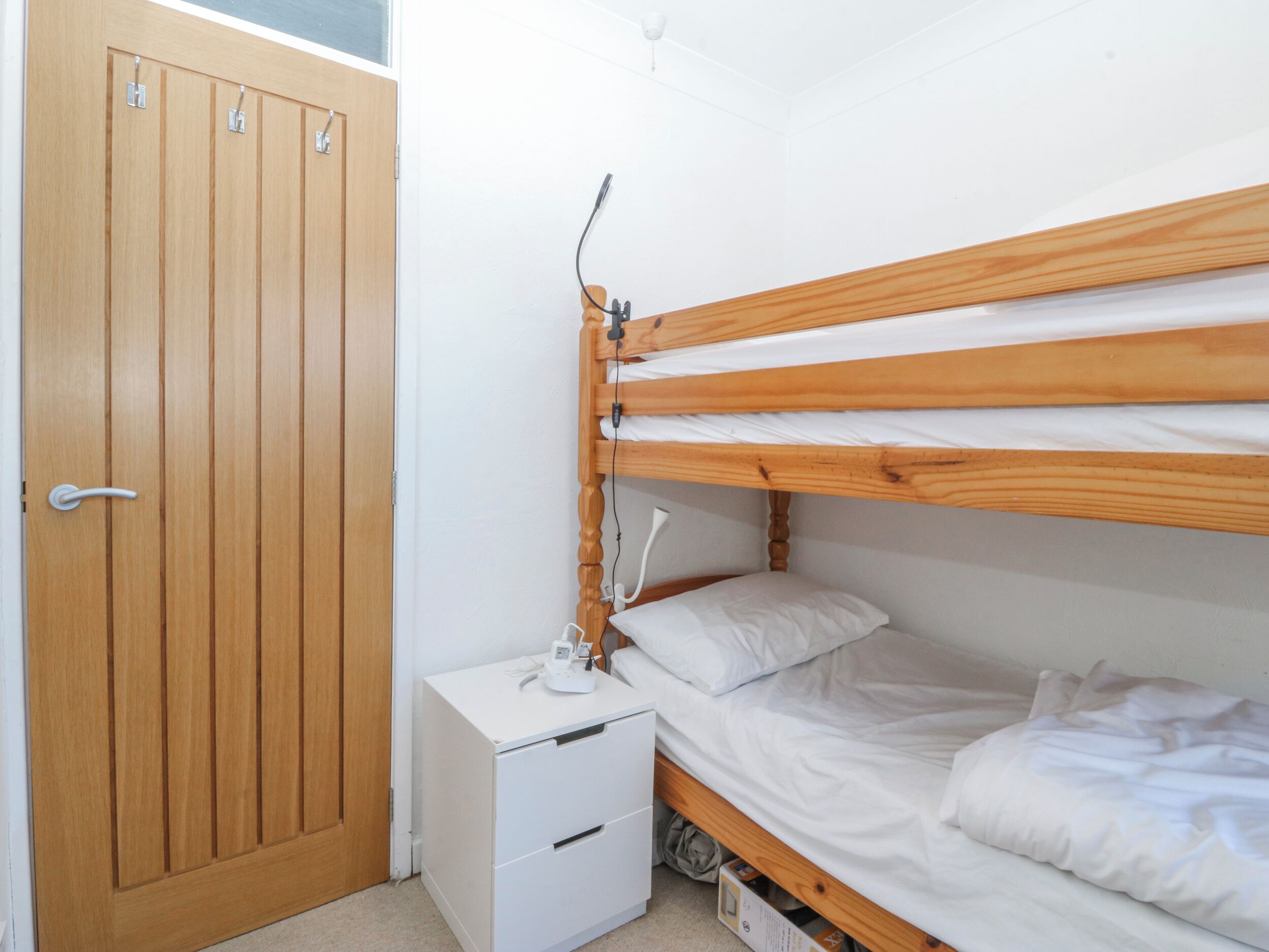 A room with a wooden door, a bunk bed, a bedside table with a lamp, and a small white cabinet.