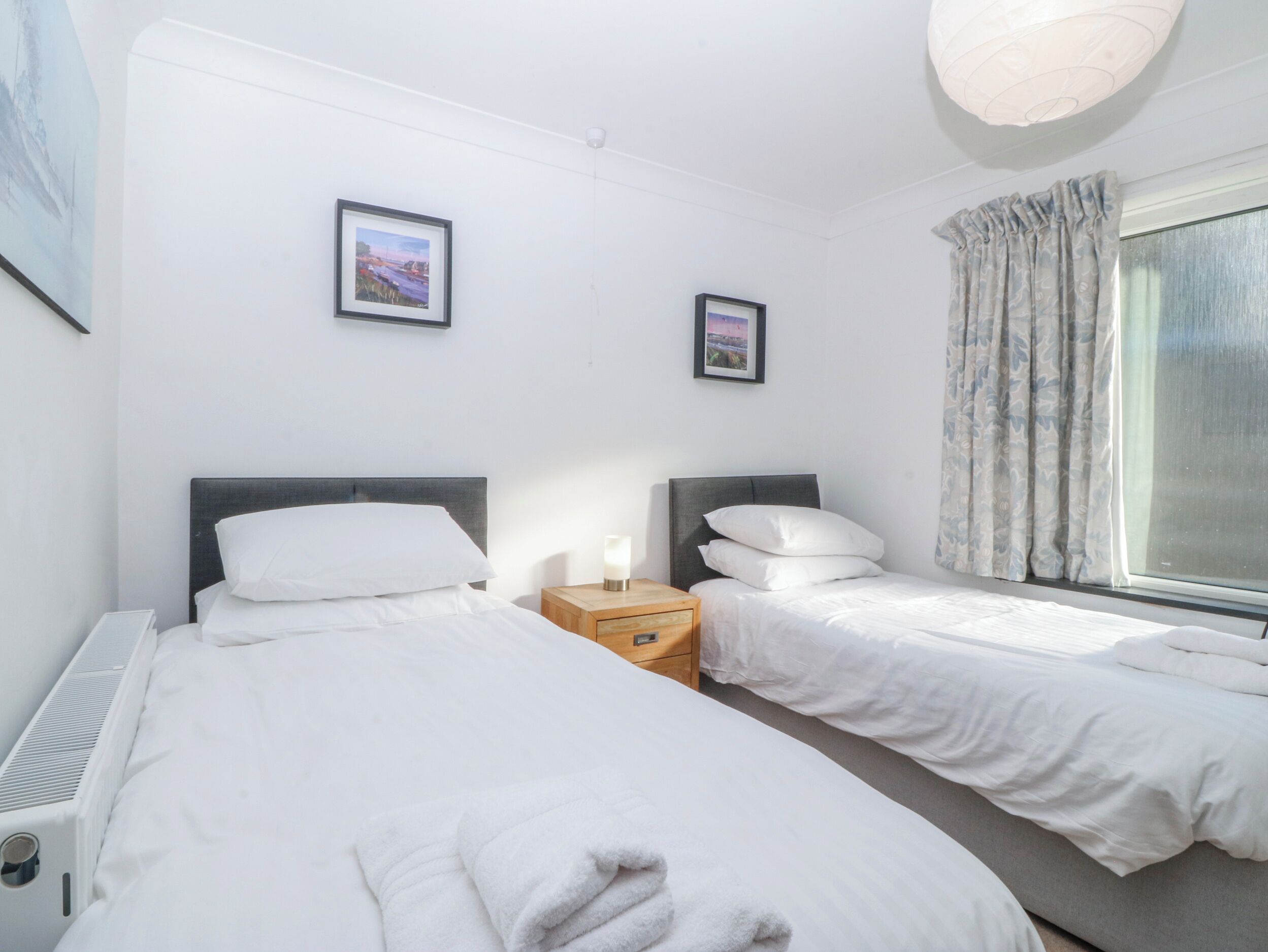 A hotel room with two beds, a bedside table with a lamp, a radiator, and a window with curtains.