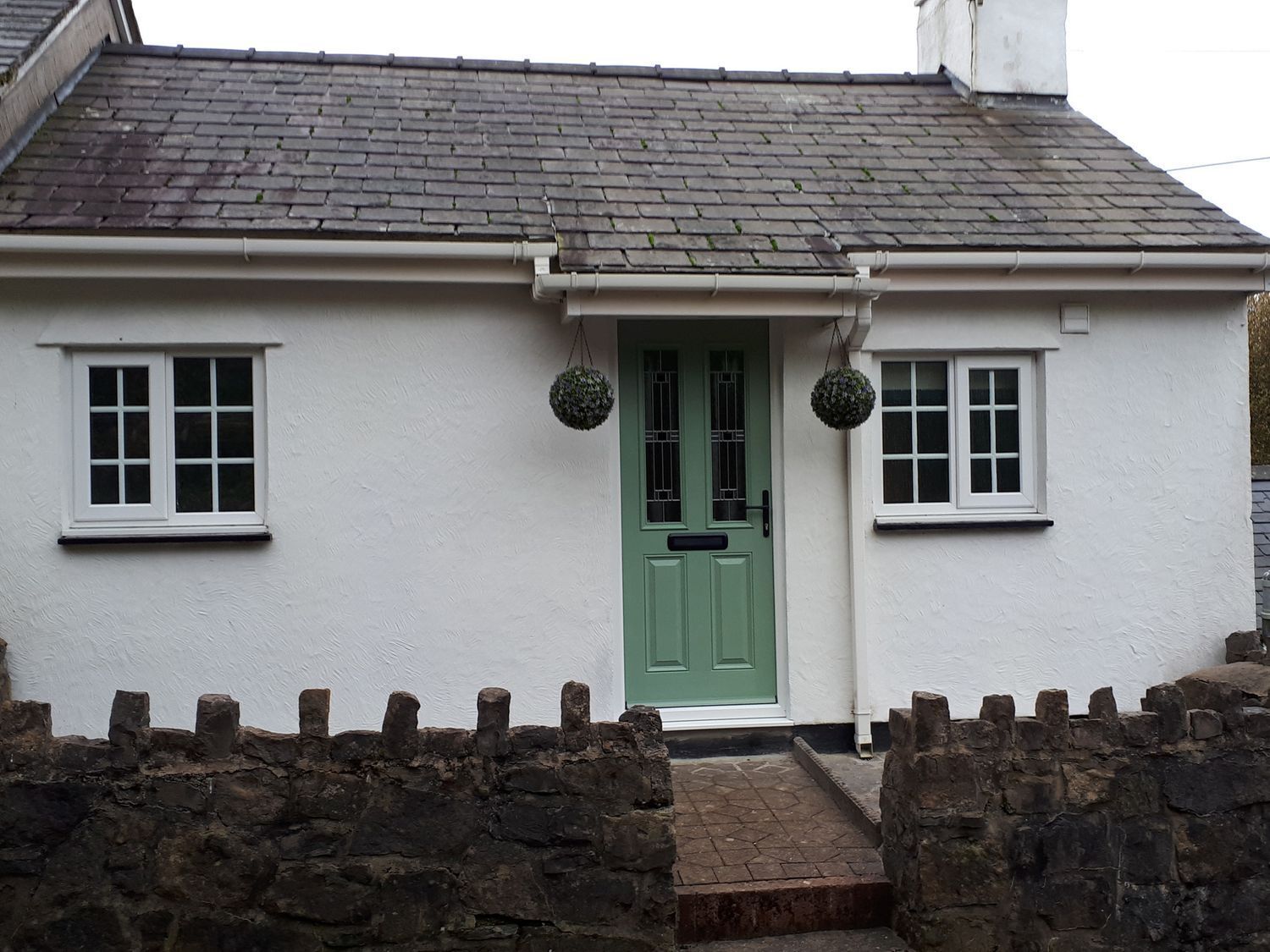 A white cottage with a green door, two windows, and a stone wall in front.