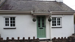A white cottage with a green door, two windows, and a stone wall in front. - Bryn Bach (Pentraeth)