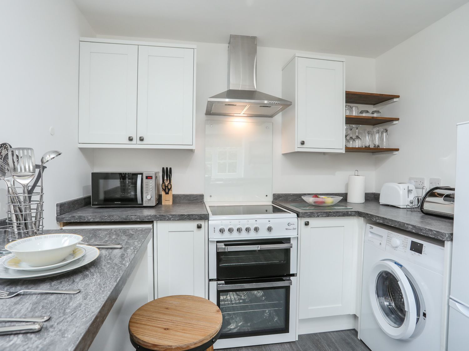 A compact kitchen with white cabinets, a microwave, a stove, a washing machine, and a wooden stool.