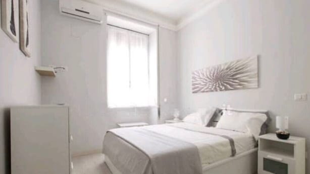 A bedroom with a bed, bedside tables, a nightstand, a wall-mounted air conditioner, and a window with curtains.