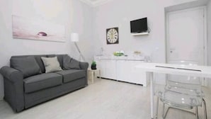 A modern living room with a grey sofa, a white dining table, and clear chairs. A small side table with a lamp and a potted plant is visible.