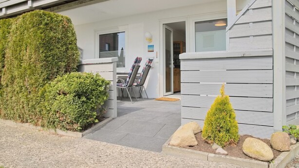 Ein modernes Gebäude mit überdachter Terrasse, einem kleinen Garten mit Steinen und einer geschnittenen Hecke sowie einer Tür zu einem Innenraum mit Stuhl und Tisch.