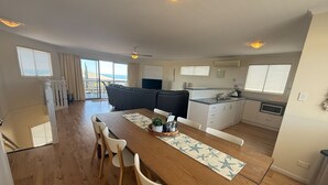 A spacious living area with a wooden dining table, white chairs, a grey sofa, and a kitchen area with white cabinets and a microwave. - Seaviews at Stansbury - Modern and spacious! Off Peak special available! (Stansbury)