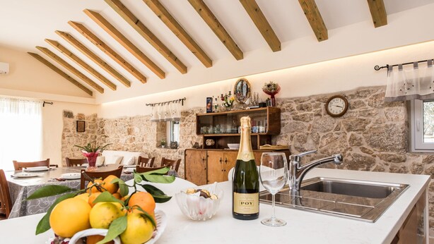A kitchen with stone walls, wooden beams, a white countertop, a sink, a bottle of wine, and a bowl of fruit. - Villa Didova kuća ZadarVillas (Podlug)