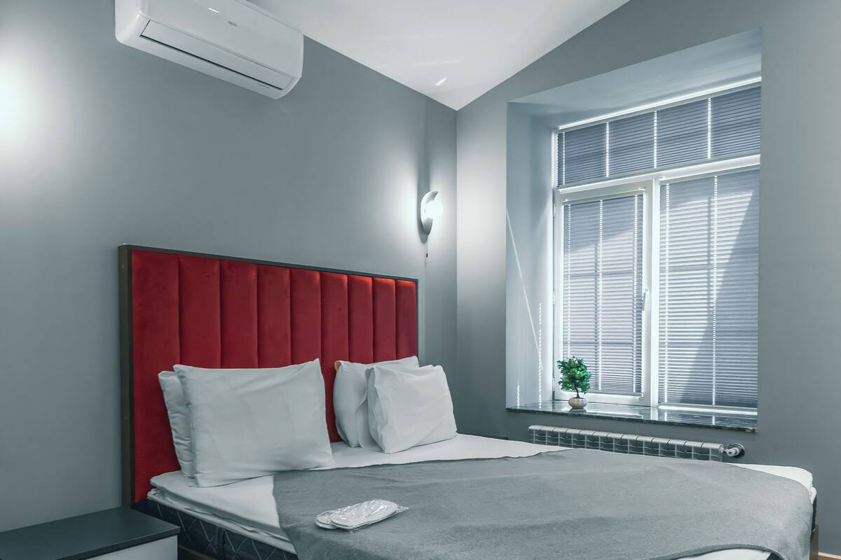 Un dormitorio moderno con cabecera roja, cama con manta gris, mesita de noche con cajones y ventana con persianas.