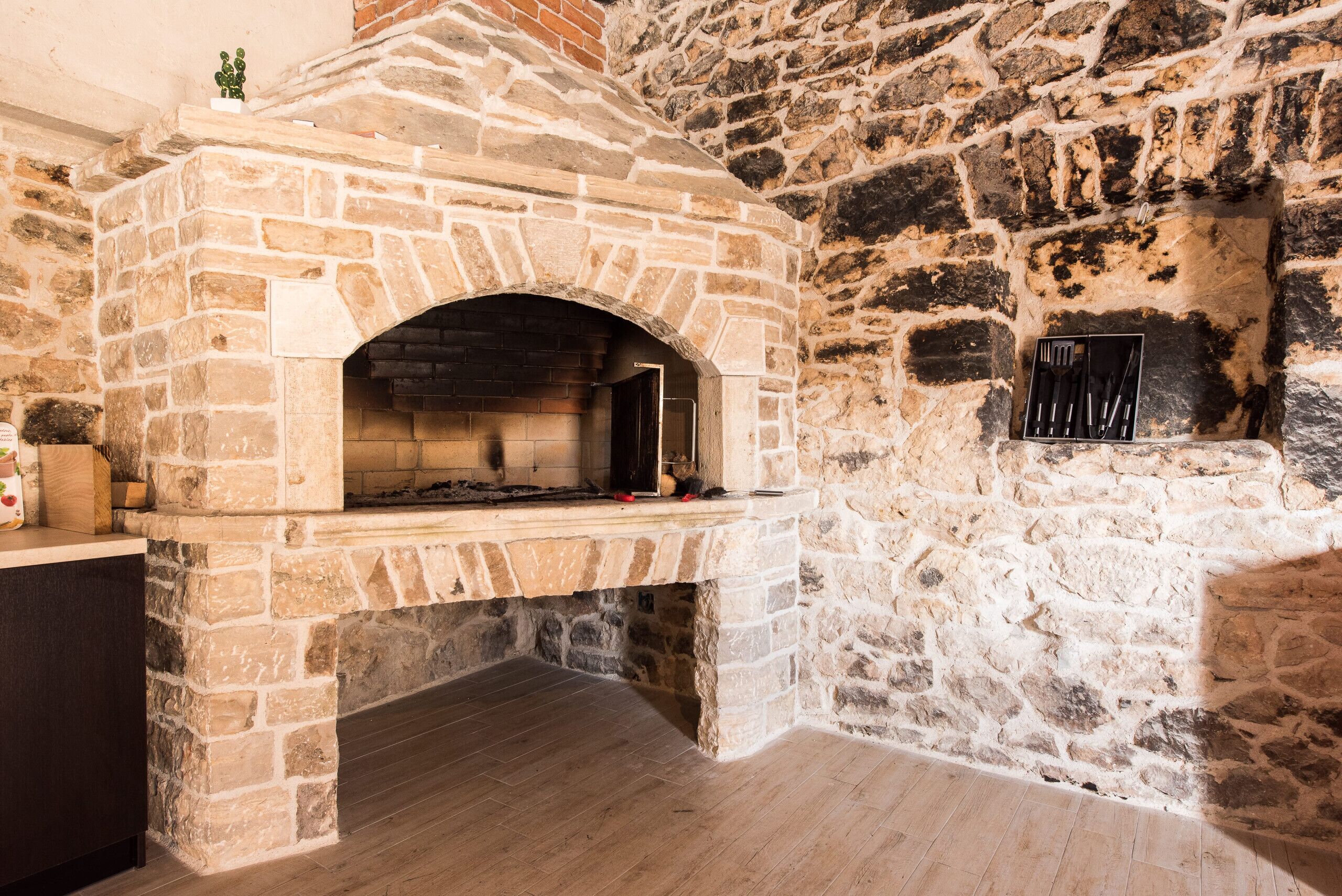 A stone-built brick oven with a shelf above it, a small plant on the shelf, and a set of utensils hanging on the wall to the right.