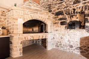 Ein aus Stein gemauerter Backofen mit Ablage darüber, eine kleine Pflanze auf der Ablage und ein Werkzeugset, das an der rechten Wand hängt.