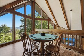 Ein runder Holztisch mit Stühlen, große Fenster und Blick auf Grünflächen und Hügel.