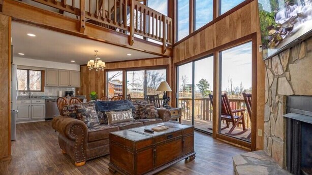 A spacious living room with a high ceiling, wooden flooring, a large sofa with a 'HOME' pillow, a wooden coffee table, and a fireplace. - 5BR Gatlinburg Family Getaway with Views, Hot Tub & Game Room in Chalet Village (Gatlinburg)