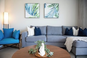 A modern living room with a grey sofa, blue cushions, a white decorative piece, and two framed leaf paintings on the wall. - Port Views On Airlie Premier (Airlie Beach)