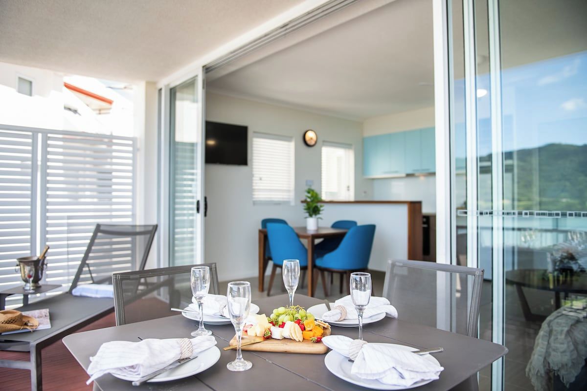 Un comedor con una mesa dispuesta para cuatro personas, un frutero y vista a la cocina y comedor a través de un ventanal.