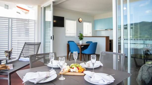 Un comedor con una mesa dispuesta para cuatro personas, un frutero y vista a la cocina y comedor a través de un ventanal.