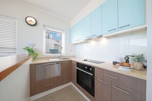 A compact kitchen with a dishwasher, oven, and a window with blinds. - Port Views On Airlie Premier (Airlie Beach)