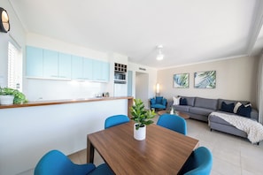 Ein modernes Wohnzimmer mit Esstisch, blauen Stühlen, einem grauen Sofa und einer Küchenzeile mit hellblauen Schränken.