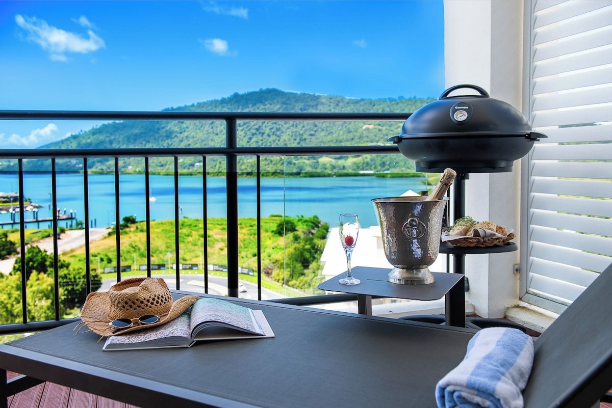 Un balcon avec vue sur un lac et des collines, un barbecue, un livre et un verre de vin.