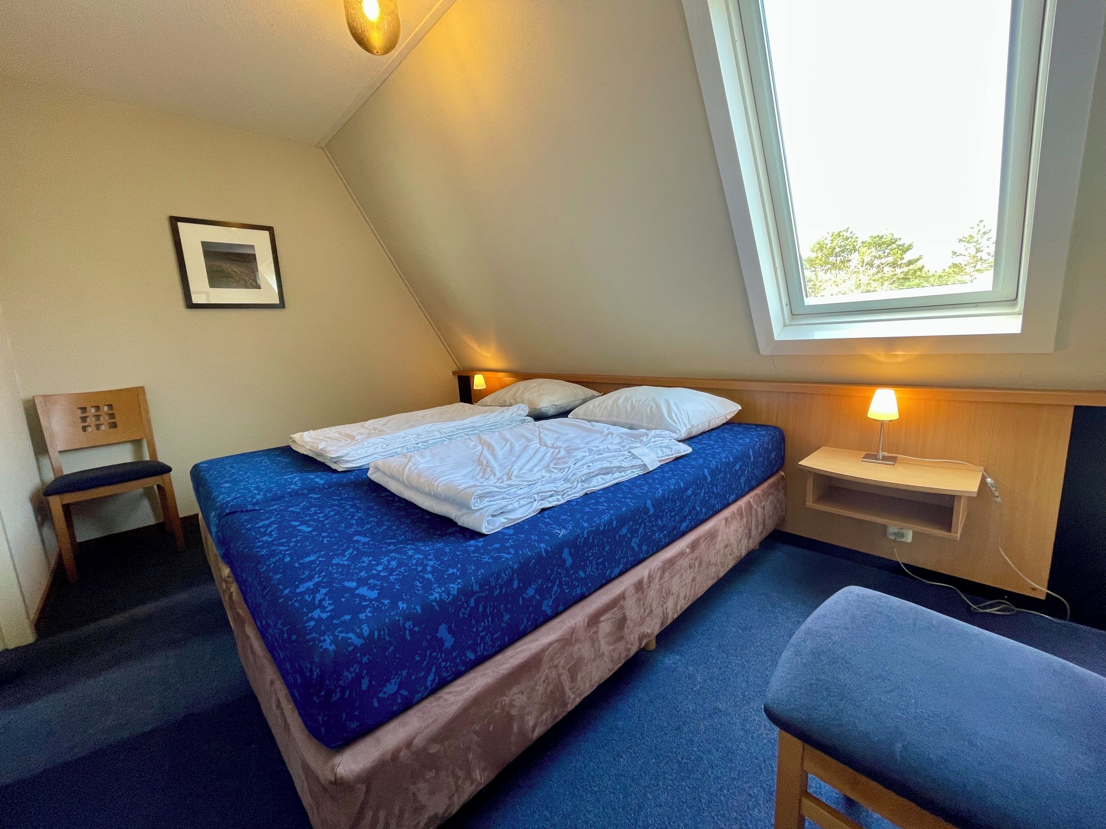 Un lit simple avec un matelas bleu, une table de chevet avec une lampe, une chaise et une fenêtre donnant sur des arbres.