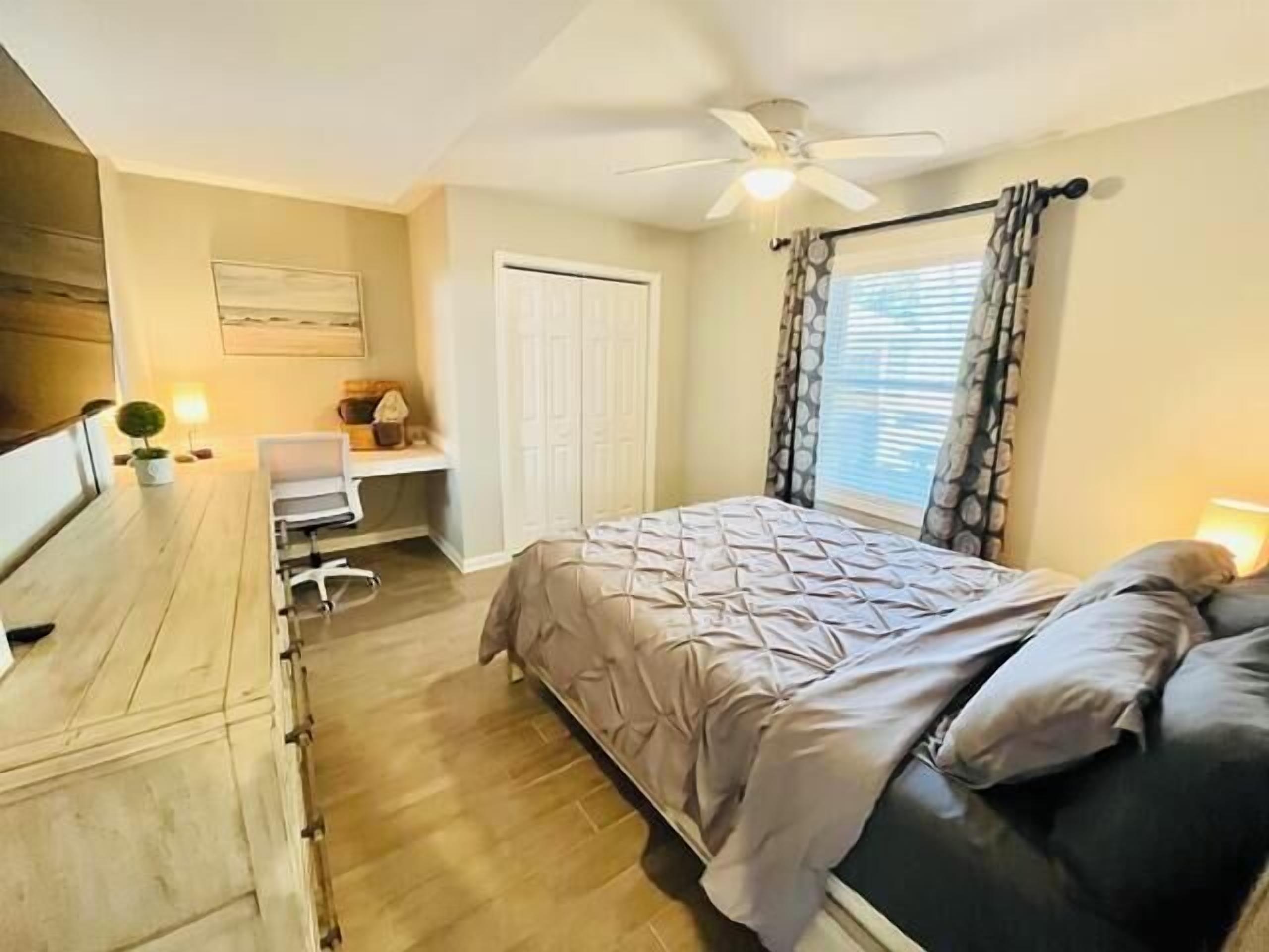 Une chambre à coucher comprenant un lit, un bureau, une chaise, un ventilateur de plafond et une fenêtre avec des rideaux.