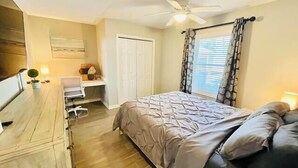 Une chambre à coucher comprenant un lit, un bureau, une chaise, un ventilateur de plafond et une fenêtre avec des rideaux.