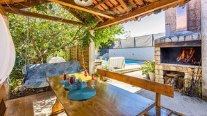 An outdoor patio with a wooden table set for dining, a stone fireplace with a fire, and a pool in the background. - Charming three-bedroom apartment near the sea (Valbandon)