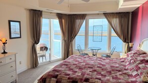 Ein Schlafzimmer mit einem großen Bett, einem Deckenventilator, Meerblick durch große Fenster und einem Balkon mit Sitzmöbeln.