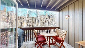 Eine überdachte Terrasse mit einem Holztisch und Stühlen, einer Glastür und Blick auf eine von Bäumen gesäumte Straße.