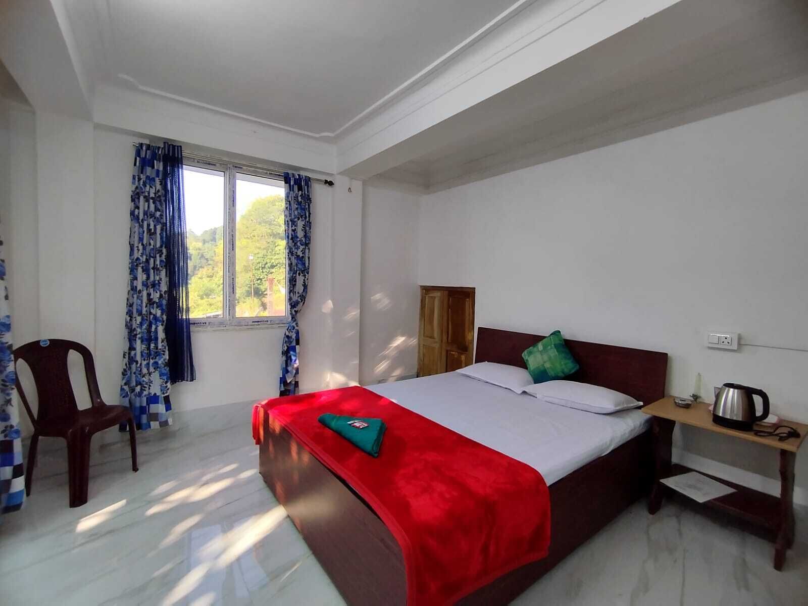 Une chambre d’hôtel avec un lit, une chaise, une fenêtre avec des rideaux, une table de chevet avec une bouilloire et une vue sur la verdure à l’extérieur.