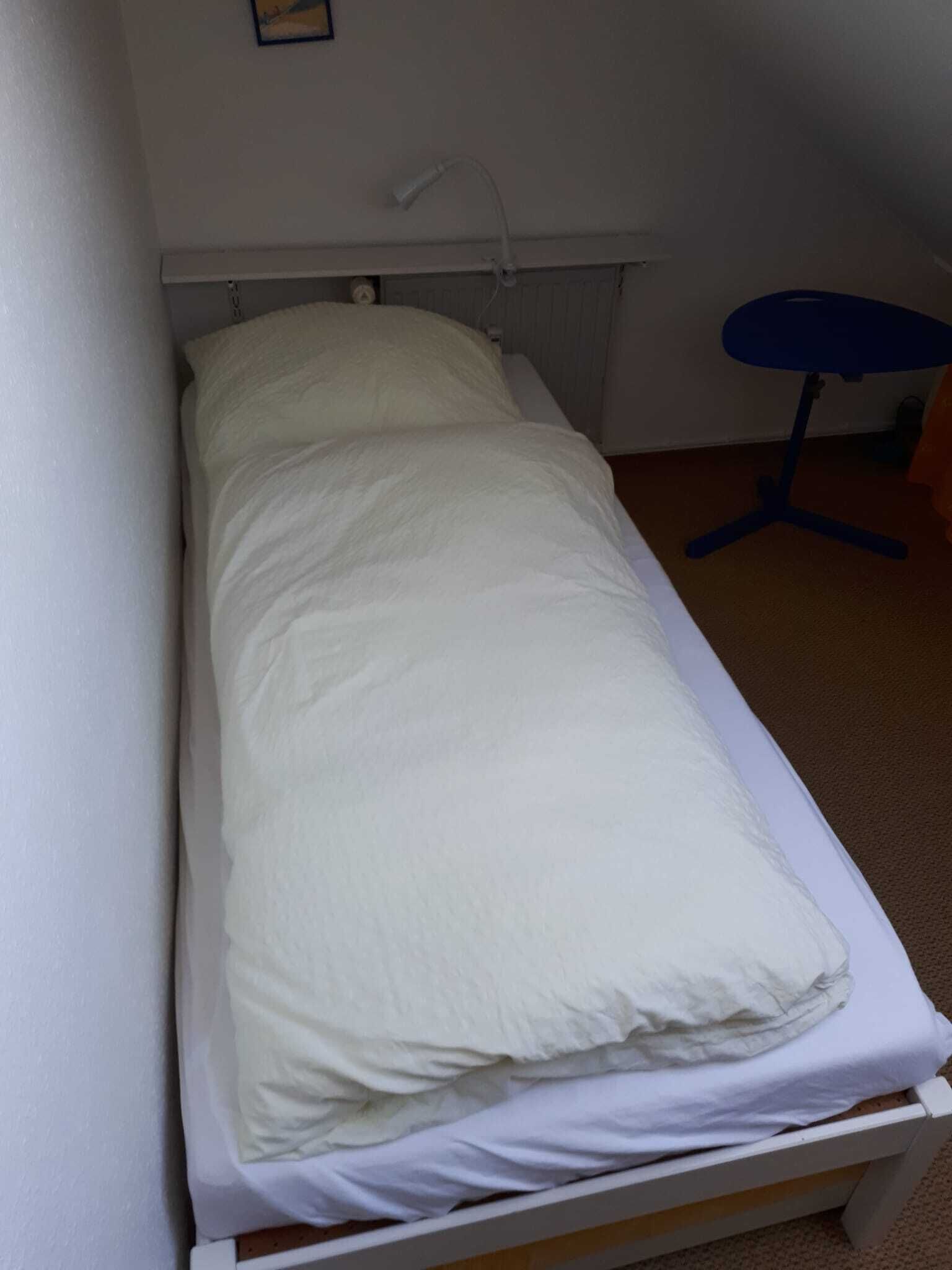 A single-bed room with a white mattress, a small bedside table, and a chair.
