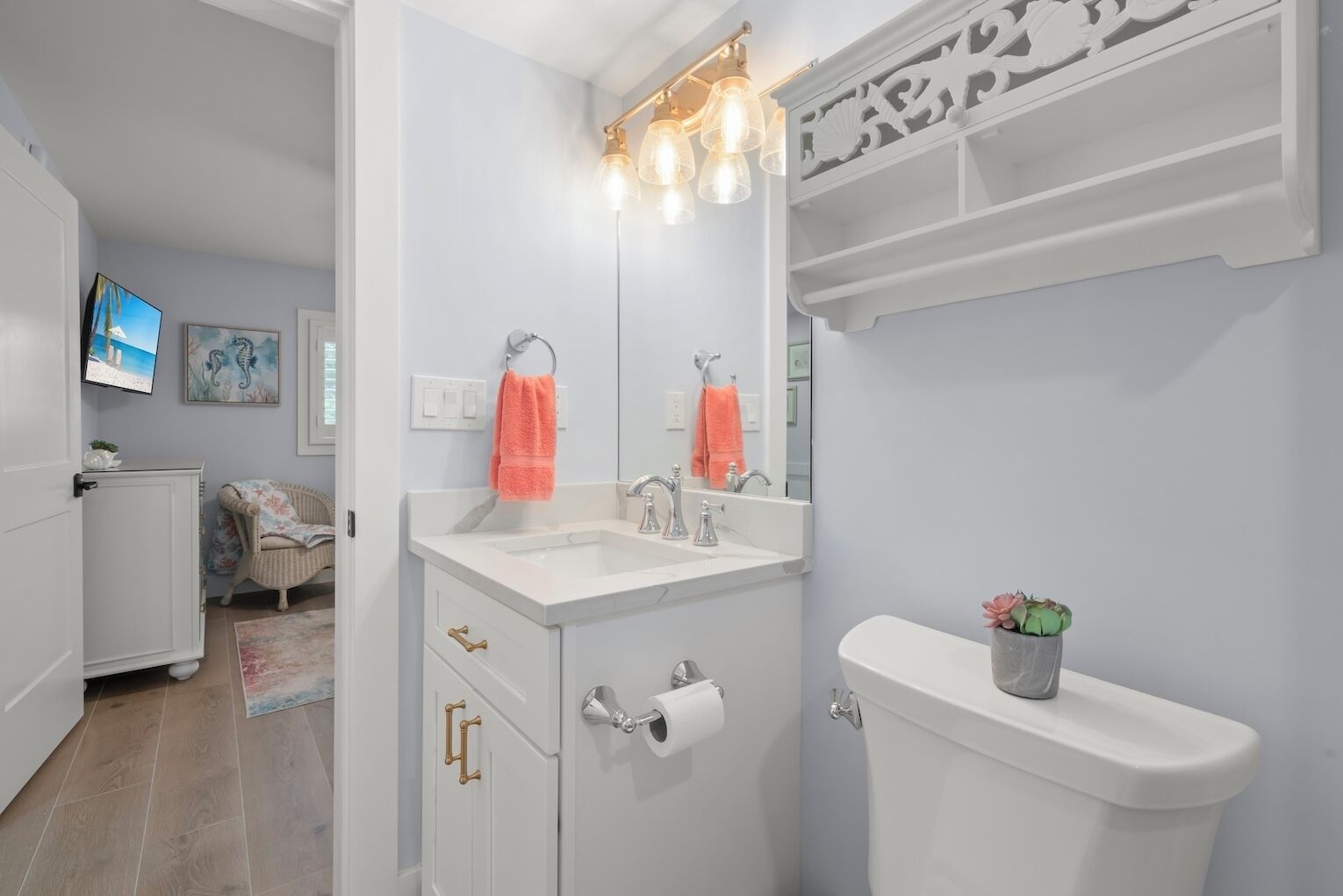 A modern bathroom with a white vanity, a toilet, and a sink with a mirror above. There are decorative lights above the sink and a small plant on the toilet tank.