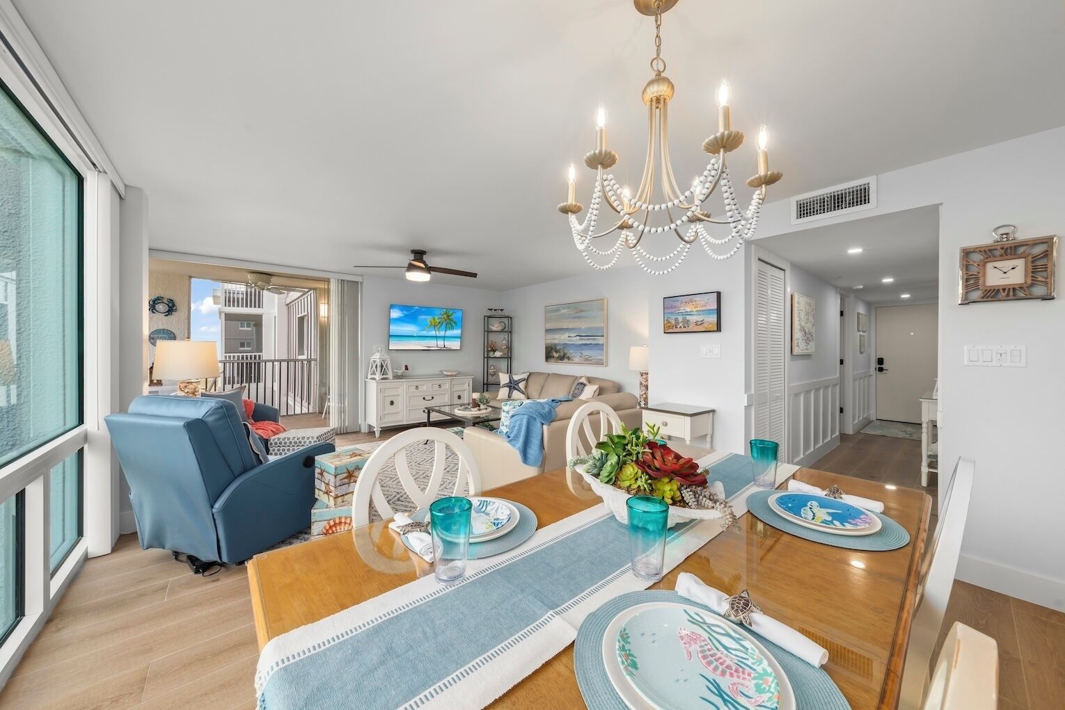 A spacious living area with a dining table set for four, a chandelier, and a balcony with a view.
