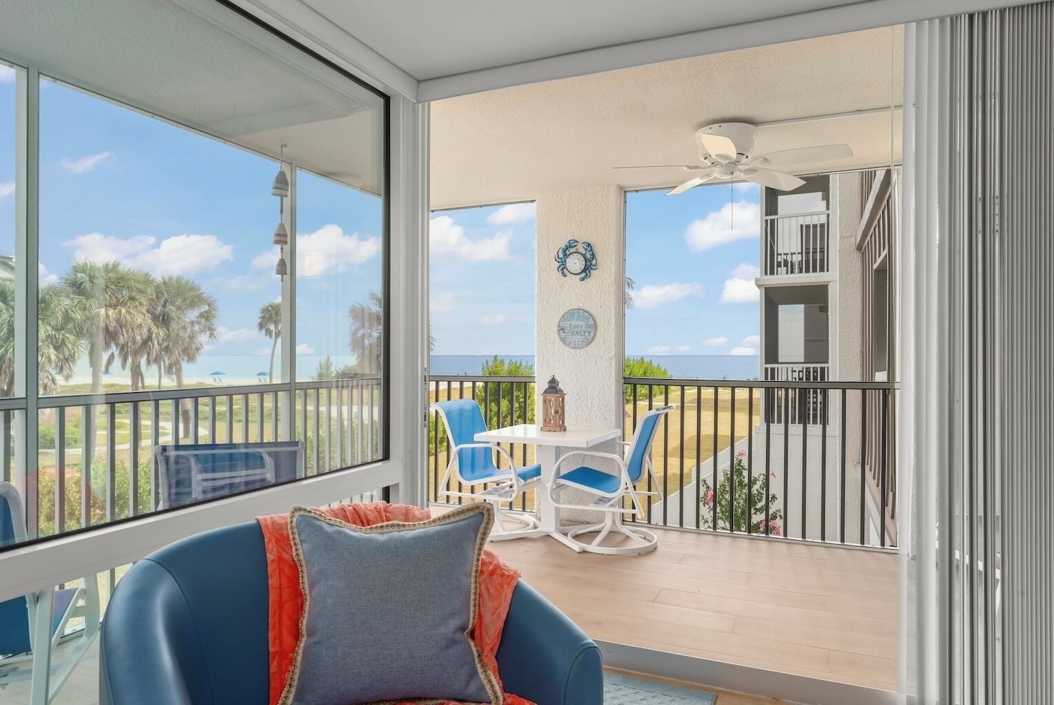 Un balcon offrant une vue sur les palmiers et l’océan, aménagé avec une table, des chaises et un fauteuil rembourré.